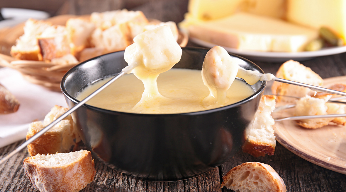 large black bowl filled with cheese surrounded by bread and being dipped into with metal skewers